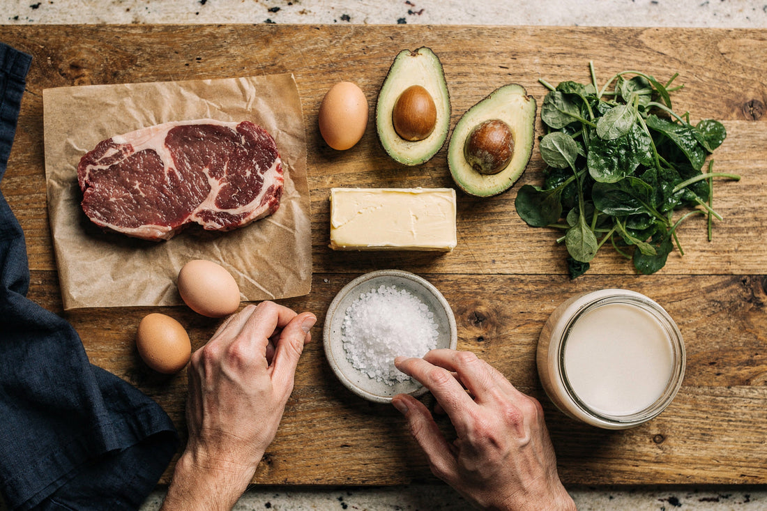 Overhead flat lay of keto foods: grass-fed ribeye, pastured eggs, butter, avocado, spinach, and sea salt on a wood table