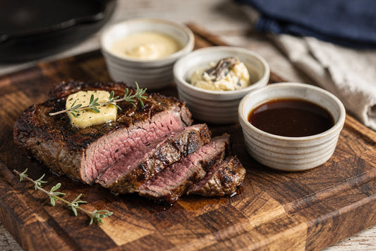 Sliced grass-fed beef steak on a wood board with small bowls of homemade tallow mayonnaise, anchovy butter, and pan sauce