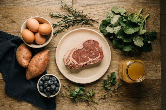 Flat lay of grass-fed ribeye, pastured eggs, sweet potatoes, leafy greens, blueberries, fresh herbs, and a jar of bone broth on a wood table