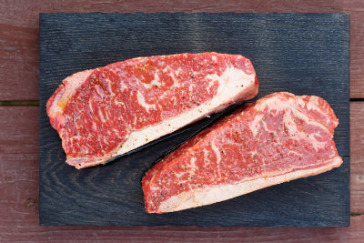 Two raw NY strip steaks on a dark slate surface with a wooden background