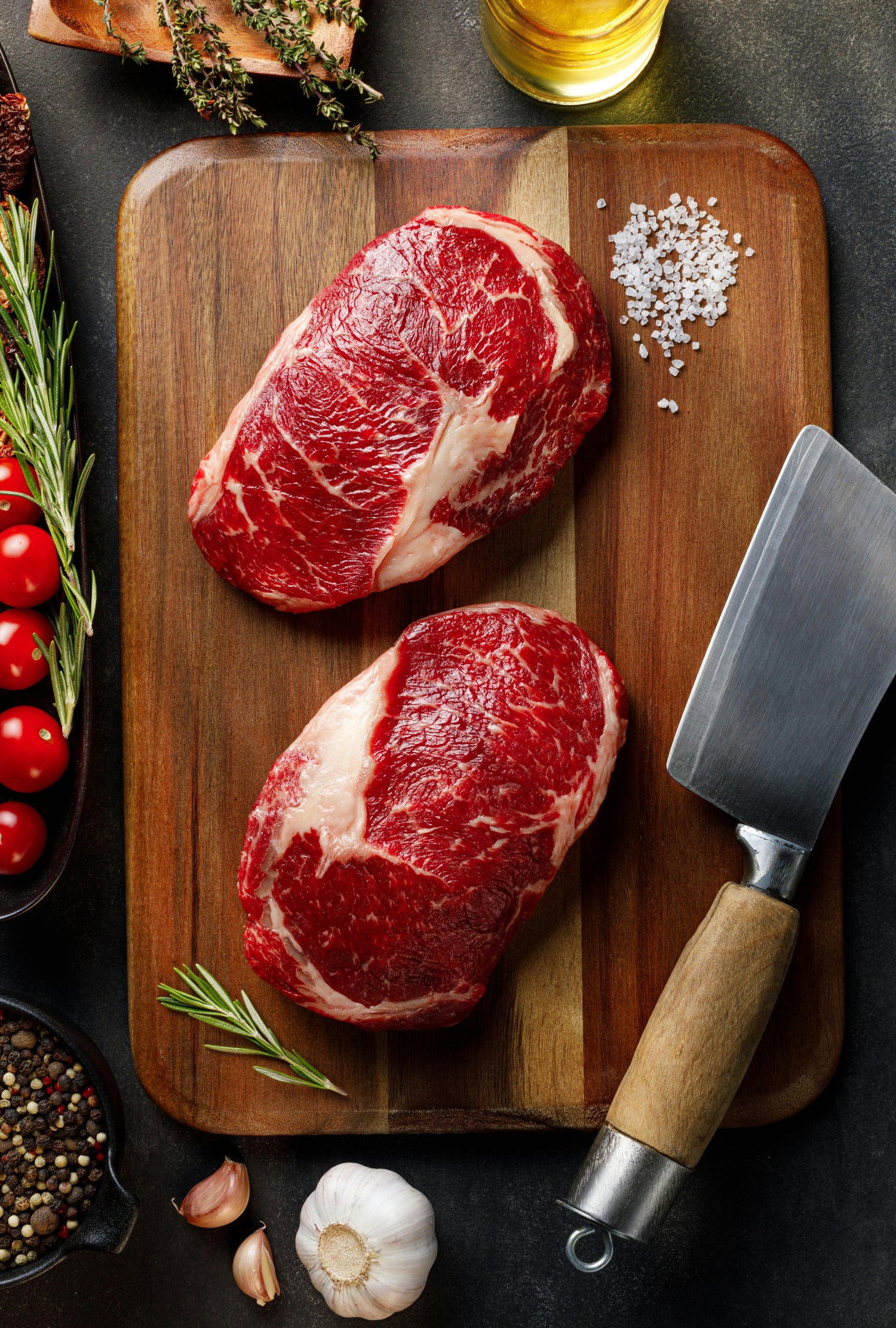 Two raw grass fed steaks on a wooden cutting board with a knife, rosemary, garlic, and tomatoes.
